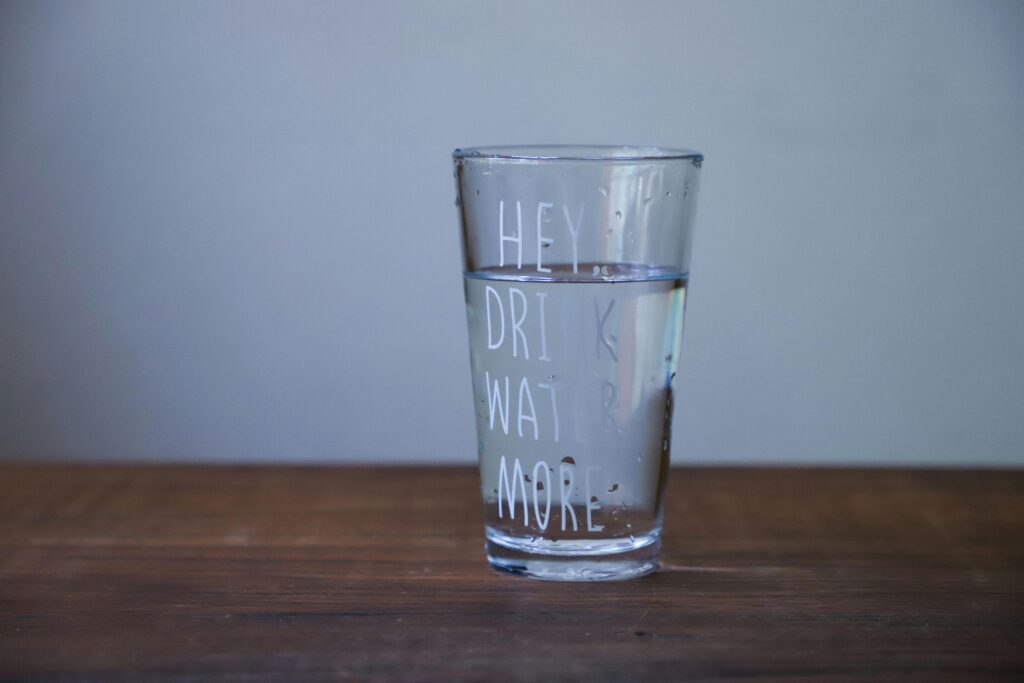 A glass of clear water on a wooden table with the text 'HEY DRINK WATER MORE'.